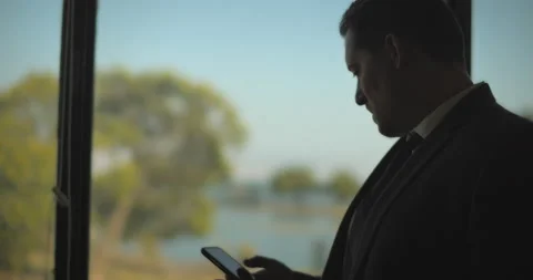 Businessman checking his phone while standing near a window and looking outsi Stock Footage 147360999