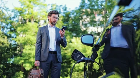 Businessman Communicating on Smartphone While Charging Electric Car Outdoors Vidéo 312865638