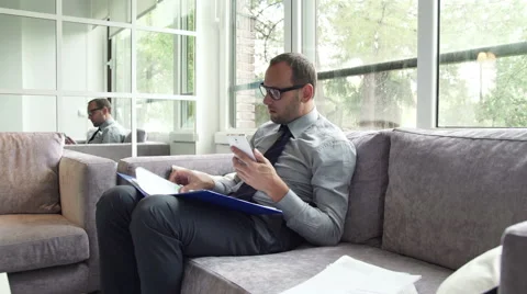 Businessman comparing data on smartphone and documents sitting on sofa HD Stock Footage 43337315