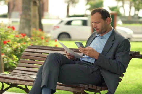 Businessman comparing data on smartphone and documents at the city bench NTSC Stock Footage 43399275
