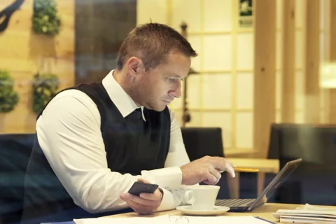 Businessman comparing data on smartphone, documents and laptop in cafe  NTSC Stock Footage 45543221