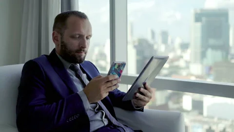 Businessman comparing data on tablet and smartphone on armchair by the window,4K Stock Footage 106556582