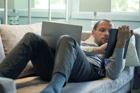 Businessman comparing data on tablet computer and laptop on sofa at home NTSC Stock Footage 43341327