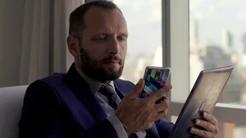 Businessman comparing data on tablet computer and smartphone on armchair by the Stock Footage 82261823