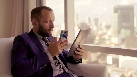 Businessman comparing data on tablet computer and smartphone on armchair by the Stock Footage 83632625