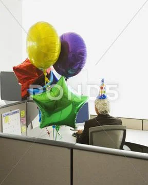 Businessman in a cubicle with a party hat on and a bunch of balloons Stock Photos