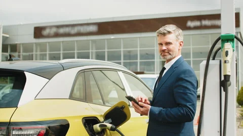 Businessman holding smartphone while charging car at electric vehicle charging Stock Footage 235220018