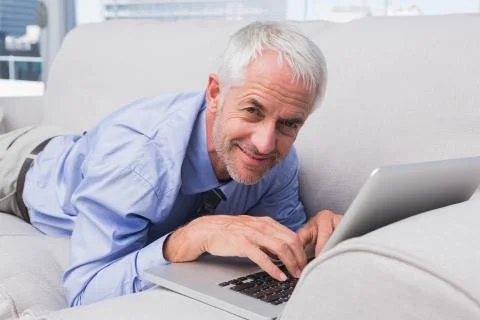 Businessman lying on couch using laptop and smiling at camera 스톡 사진