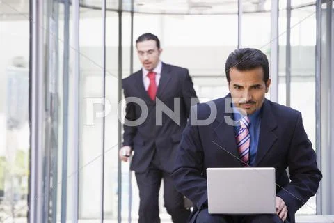 Businessman outdoors in front of building using laptop with businessman in Stock Photos