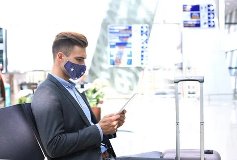 Businessman in protective mask with suitcase using tablet in hall of airport. Stock Photos