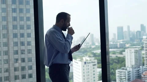 Businessman reading something on tablet computer and drinking coffee by window Stock Footage 60126239