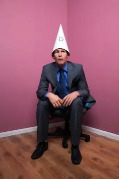Businessman sat in corner wearing dunce hat Stock Photos