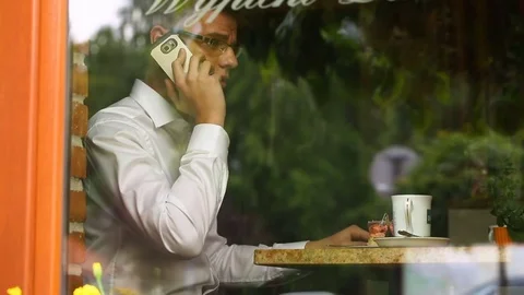 Businessman sitting behind cafe's window and looks angry while speaking on phone 스톡 동영상 80425059