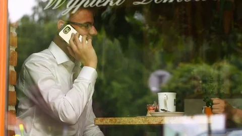 Businessman sitting behind cafe's window and chatting on cellphone Stock Footage 80425374