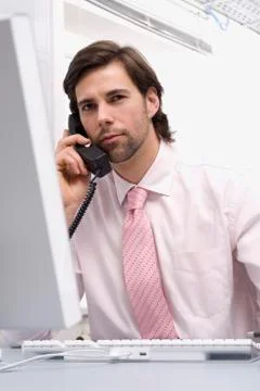 Businessman sitting by computer, using landline phone, close-up Stock Photos