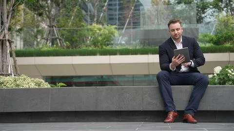 Businessman sitting at park while working on tablet by reading report. Urbane Stock Photos