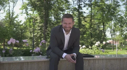 Businessman smiling to camera sitting on wall in park. 库存影片 52784697