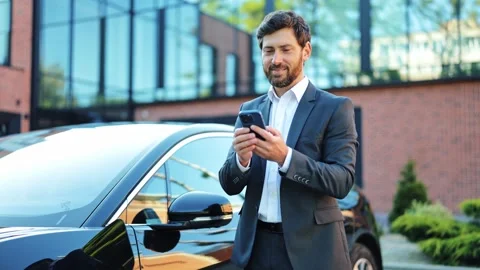 Businessman Smiling While Using Smartphone Next to Charging Electric Vehicle. Stock-Footage 312865954