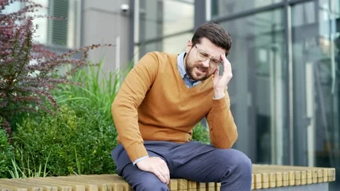 Businessman suffering from headache while sitting on a bench on street near  Stock Footage 308101107