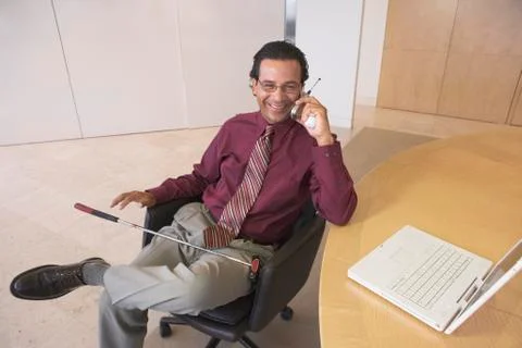 Businessman talking on cell phone in office with golf club in lap Stock Photos
