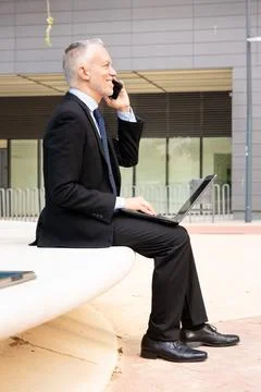 Businessman talking on his smartphone sitting with his laptop outdoors. Stock Photos