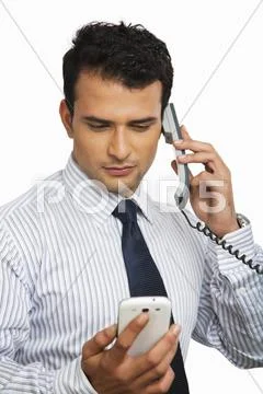 Businessman talking on a landline phone while looking at a mobile Stock Photos