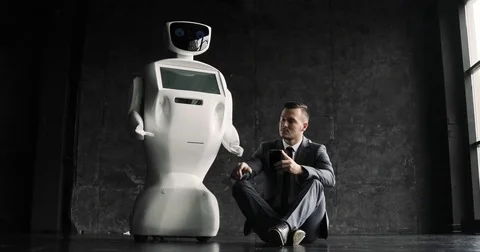 Businessman uses a smartphone while sitting on the floor next to a robot. Modern Stock-Footage 80418624