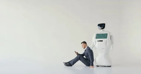 Businessman uses a smartphone while sitting on the floor next to a robot. Modern Stock Footage 80422804