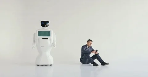 Businessman uses a smartphone while sitting on the floor next to a robot. Modern Stock Footage 80423494