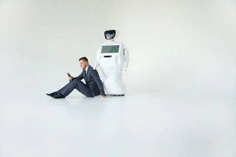 Businessman uses a smartphone while sitting on the floor next to a robot. Modern Stock Photos