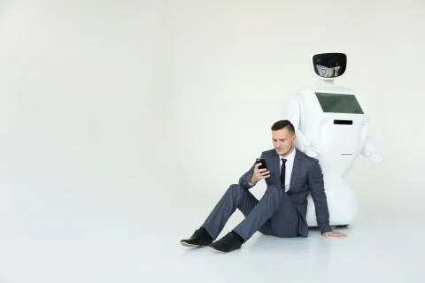Businessman uses a smartphone while sitting on the floor next to a robot. Modern Stock Photos