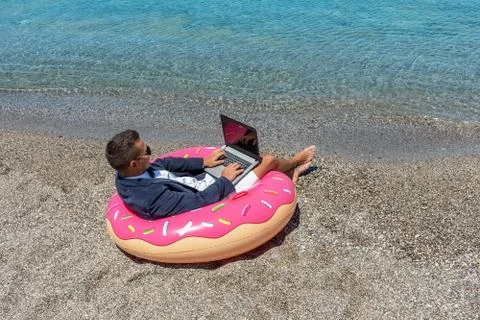 Businessman using laptop computer on inflatable donut on tropical beach. Stock Photos