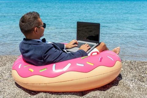 Businessman using laptop computer on inflatable donut on tropical beach. Stock Photos