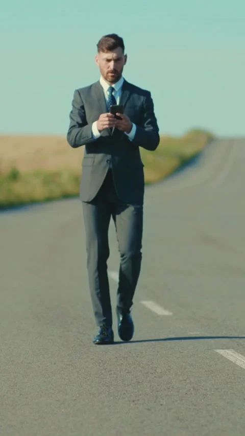 A businessman is walking alone on an empty road, casually holding a phone in his Stock Footage 316538404