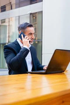Businessman working with laptop while talking on smartphone outdoors. Stock-Fotos