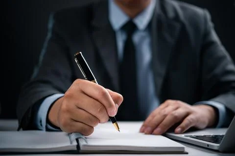 Businessman writing notes in a notebook while working on a laptop at a desk.. Stock Photos
