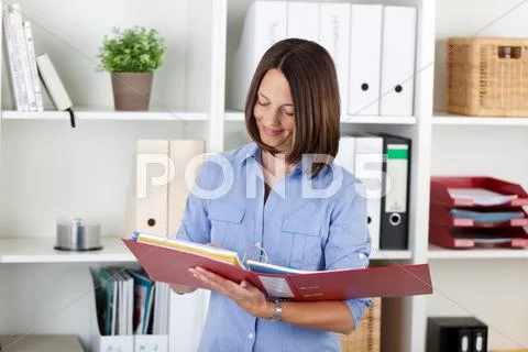 Businesswoman reading documents in binder against shelves Stock Photos