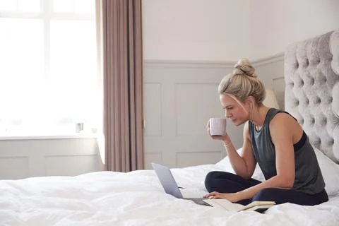 Businesswoman Sitting On Bed With Laptop Working From Home With Hot Drink During Stock Photos