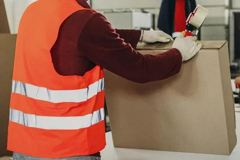 In a bustling warehouse, a dedicated worker fastens a cardboard box with tape Stock Photos