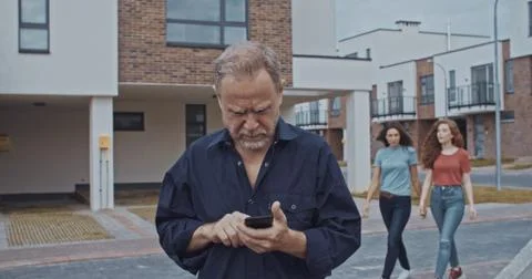 Busy elder man using smartphone outdoor asking for help of pretty young girls Stock Photos