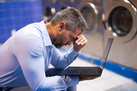 Busy man doing laundry and working on laptop. Stressed employee multitasking Stock Photos