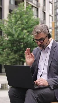 Busy man using headset while talking online with laptop outdoors. Stock Photos