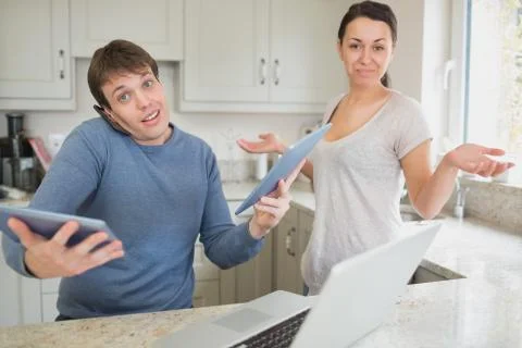 Busy man using two tablets and laptop with wife holding hands up questioning Stock Photos