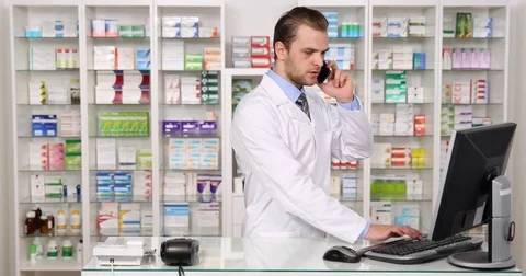 Busy Pharmacist Man Talking Mobile Phone Writing on Computer in Pharmacy Store Stock Footage 77003608