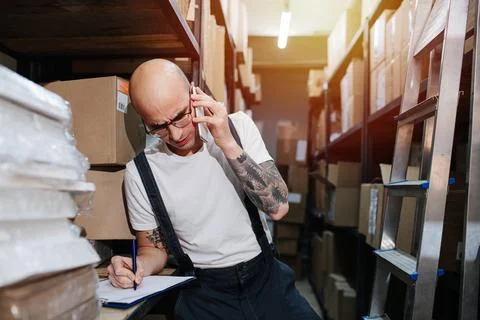 Busy warehouse worker writing down list of things, talking on the phone Stock Photos