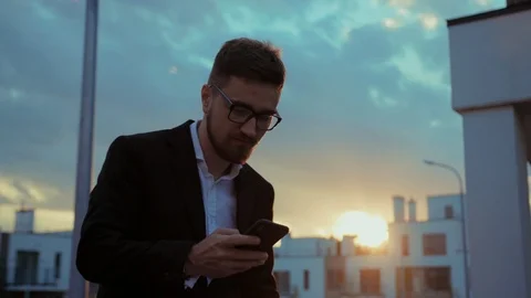 Busy young enterpreneur in suit using mobile phone at lovely sunset in city Stock Footage 113799899