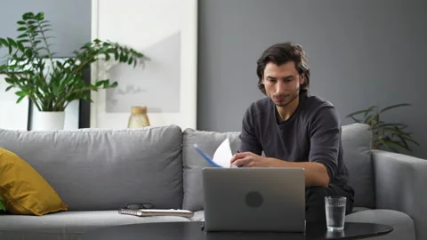 Busy young man doing paperwork at home using laptop sitting on the sofa Stock Footage 149259717