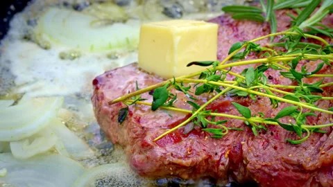 Butter cube melting on seared steak while thyme and sliced onions bubble in pan Stock Footage 325839875