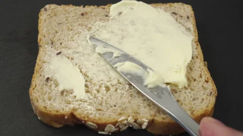 Butter is spread with a knife over a slice of cereal bread on a gray stone su Stock Footage 136167607