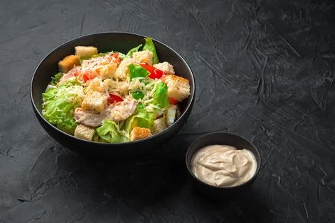 Caesar salad in a black ceramic plate with sauce on a black background. Stock Photos